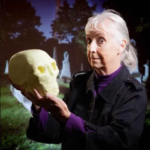 An elder with white hair posing in front of a cemetary scene and holding a skull